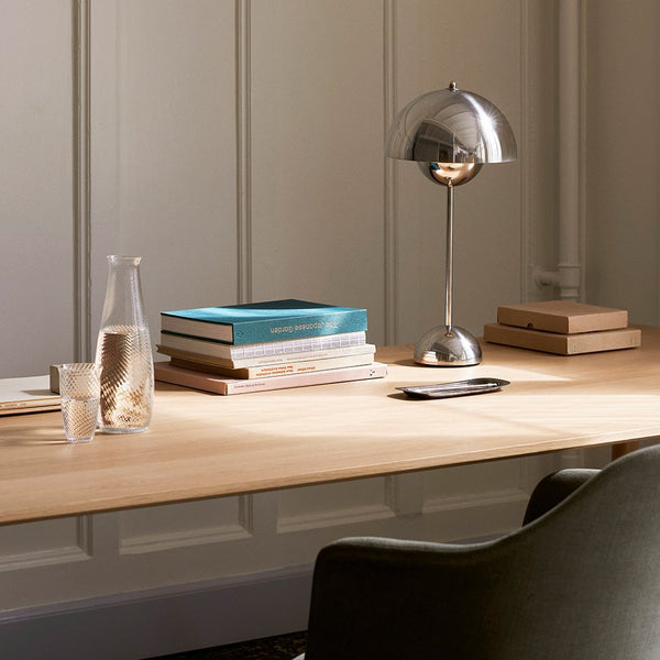 Minimalist desk with natural wood tones, silver lamp, stacked books in muted colors, and glassware. Elegant, modern design.