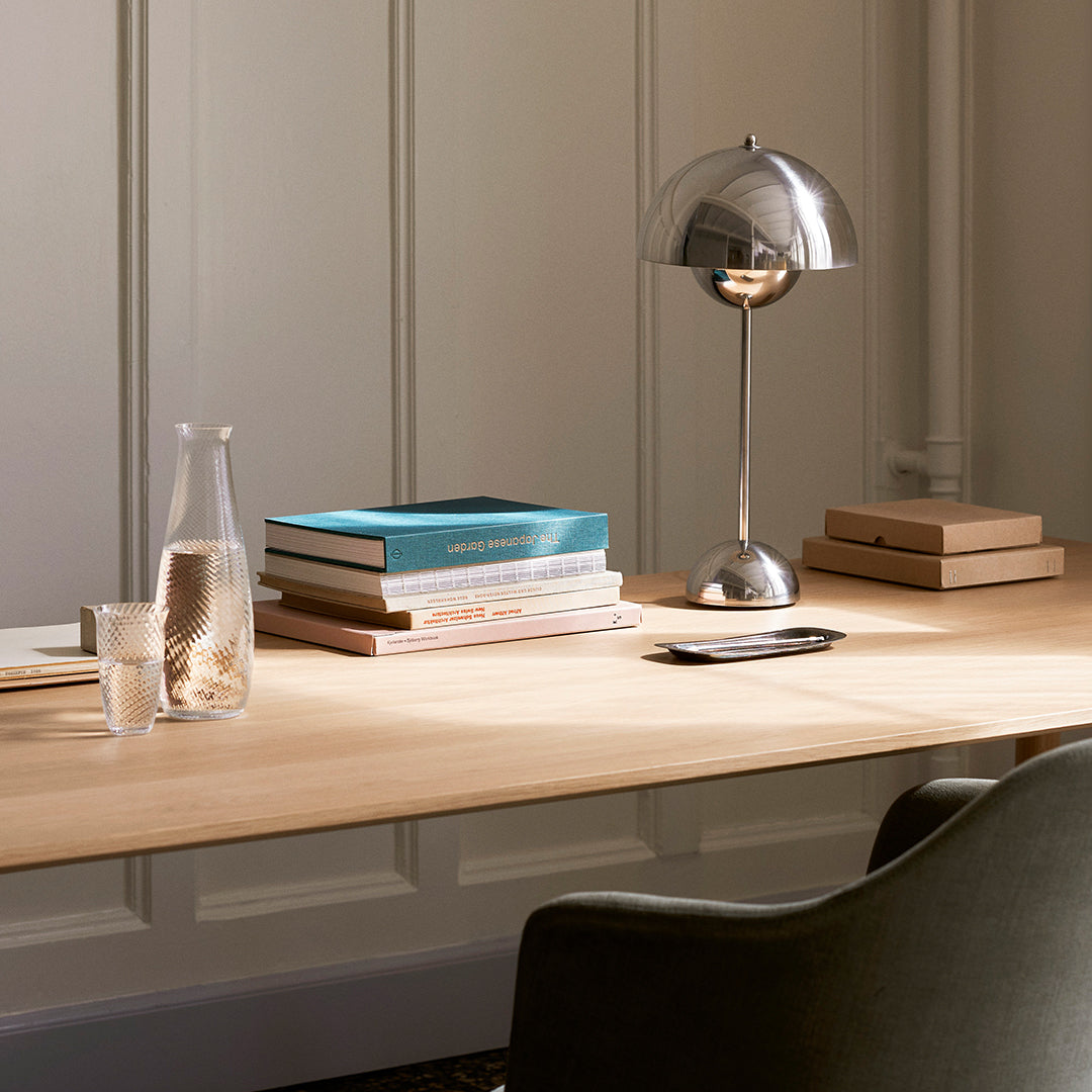 Wooden desk with a light natural finish, silver lamp, and colorful books in blue and beige tones. Clean, modern aesthetic.