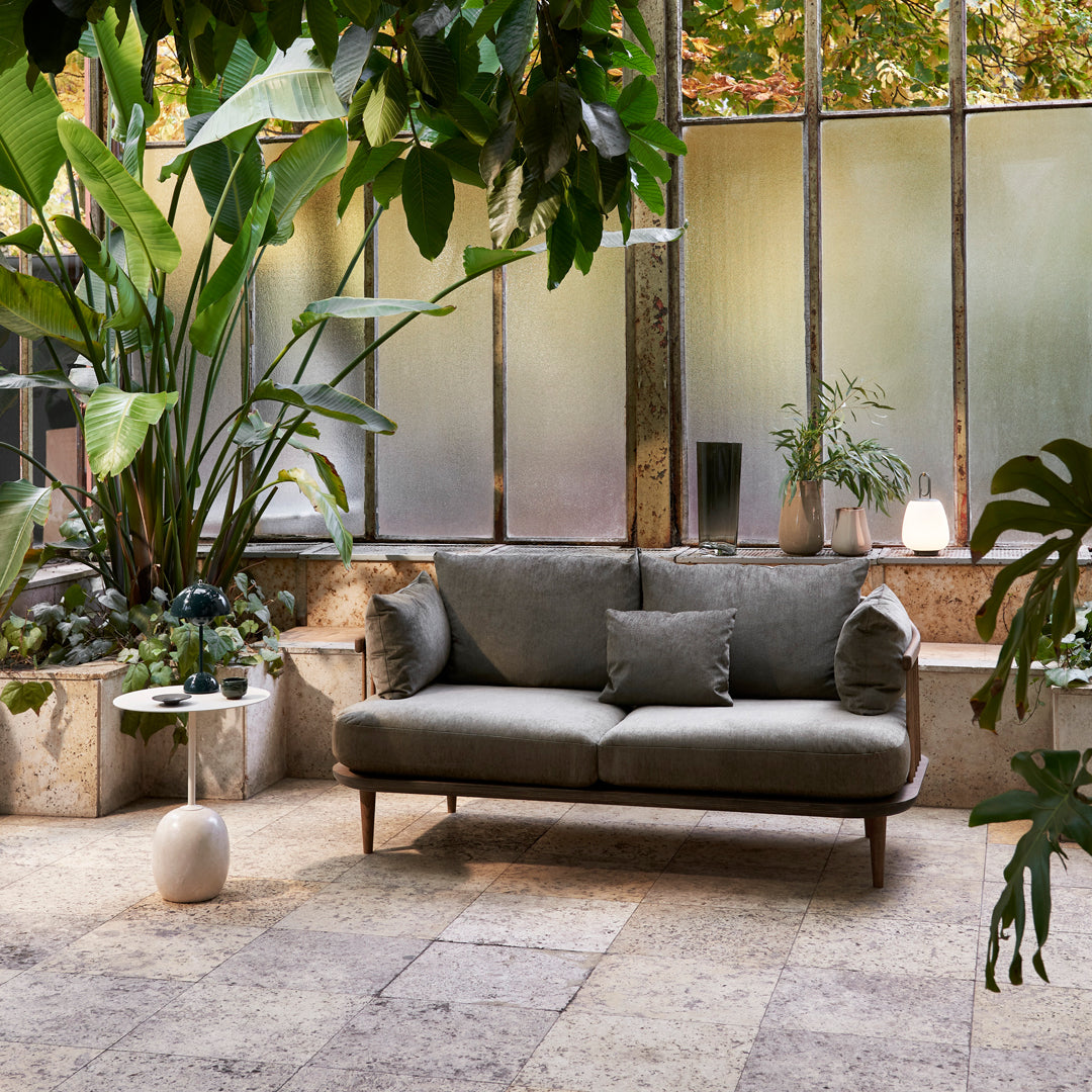 Gray upholstered sofa with textured pillows, surrounded by lush green plants and natural stone flooring.