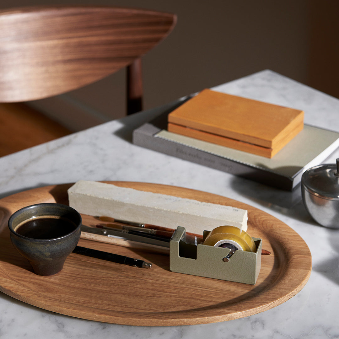 Wooden tray with dark earthy tones, bright orange and gray stationery, and a black ceramic cup on a marble surface.