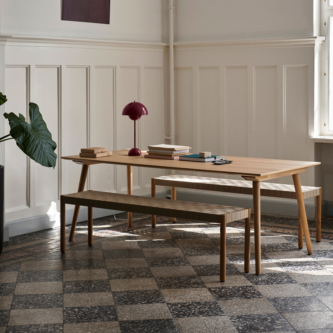 Light wooden dining table and bench with a checkered floor pattern, featuring a deep red lamp and plant accents.