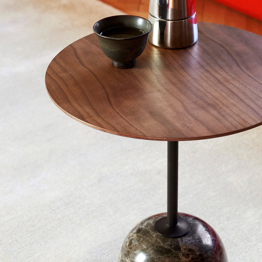 Round wooden table with rich brown grain, black metal stand, and textured stone base against a light neutral backdrop.