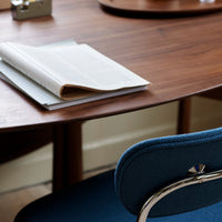 Curved walnut table with a textured dark blue chair, featuring a sleek chrome accent and layered papers.