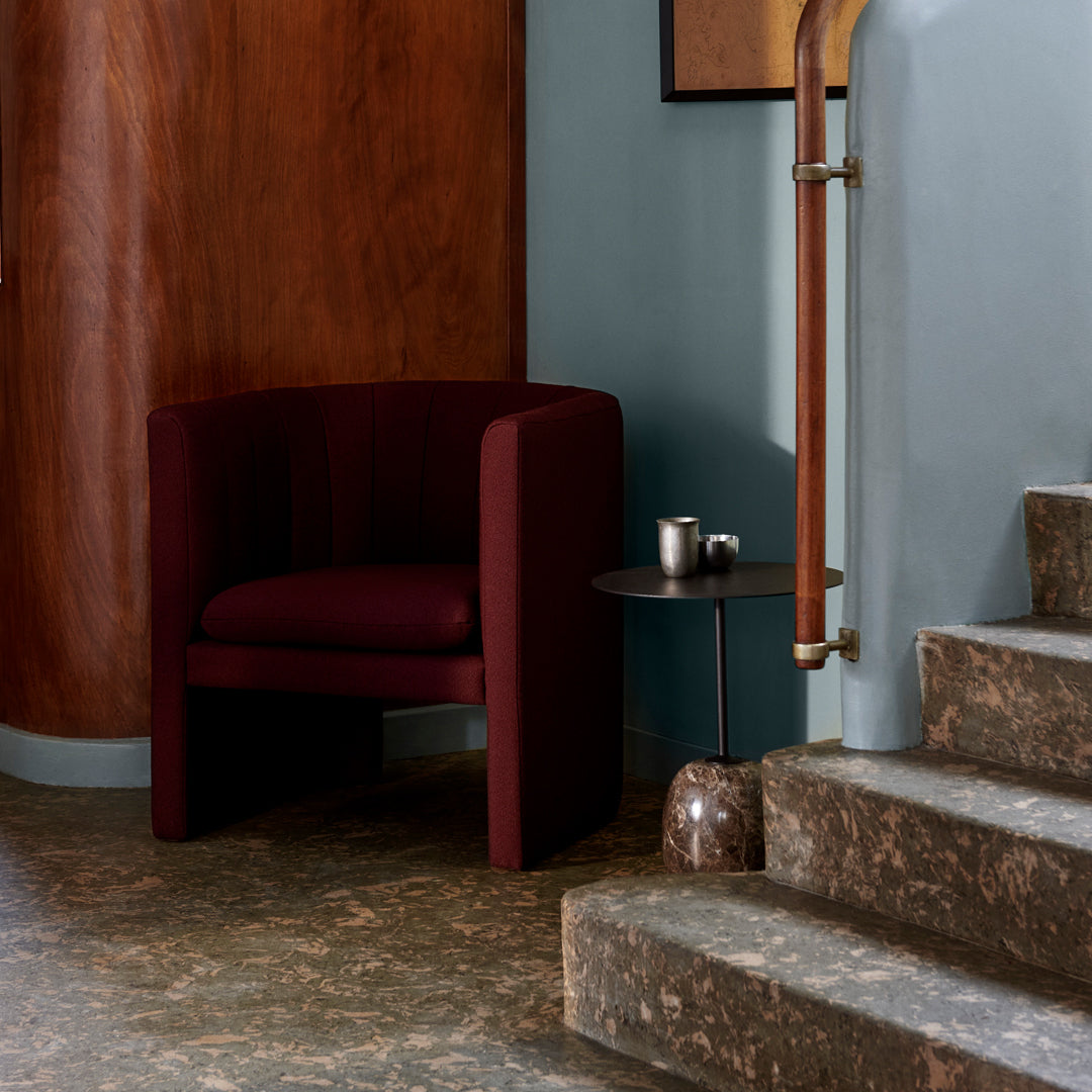 Deep burgundy accent chair with smooth upholstery, set against a warm wood wall and cool blue backdrop. Abstract patterned flooring.