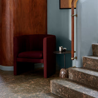 Deep burgundy accent chair with smooth upholstery, set against a warm wood wall and cool blue backdrop. Abstract patterned flooring.