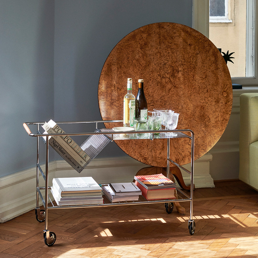 "Modern wheeled bar cart with clear glass top and silver metal frame, paired with a round, warm wood accent table."