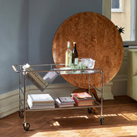 "Modern wheeled bar cart with clear glass top and silver metal frame, paired with a round, warm wood accent table."
