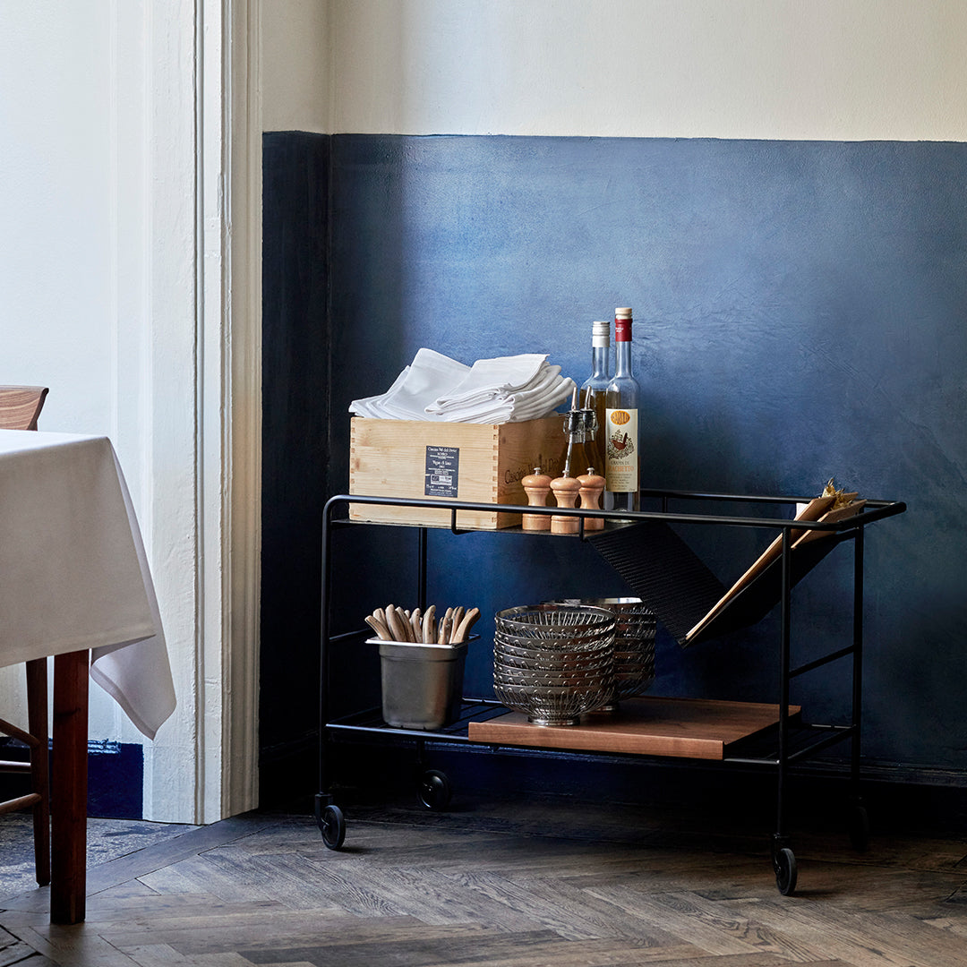 Black metal cart with wooden shelves, set against a deep blue wall, featuring white napkins and glass bottles.