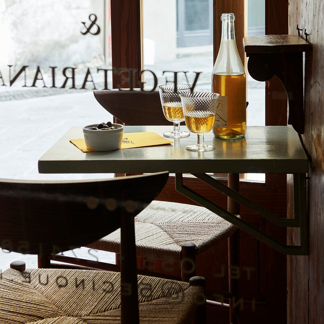 "Light green table with yellow accents and patterned seat; two glasses of golden drink by the window."