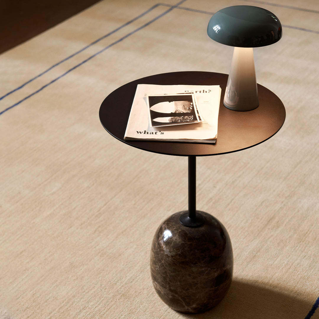 "Round black side table with a stone base, featuring a green and white lamp on a beige carpet with blue lines."