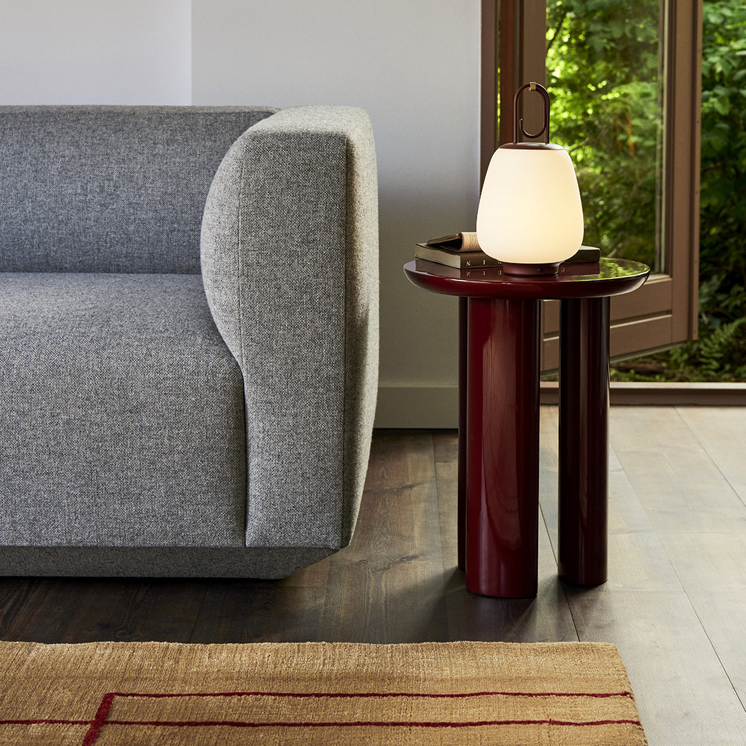 Gray fabric sofa beside a glossy red side table, with a soft white lamp on top and a textured beige rug underneath.