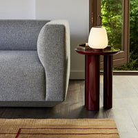 Gray fabric sofa beside a glossy red side table, with a soft white lamp on top and a textured beige rug underneath.