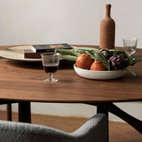 "Round wooden table with a rich brown finish, featuring a neutral plate of fruits and a textured ceramic vase."