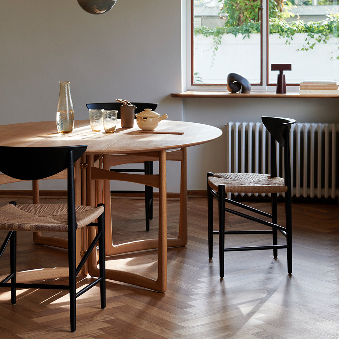 Light wood dining table with black chairs, woven seat patterns, and warm wooden flooring, complemented by natural light.
