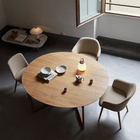 Round wooden dining table with light beige chairs in a minimalistic space, featuring simple, neutral patterns.