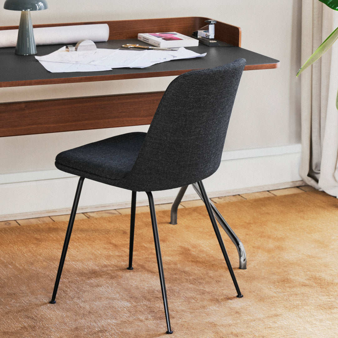 Modern black upholstered chair with sleek steel legs, set against a warm, neutral-toned floor and desk backdrop.