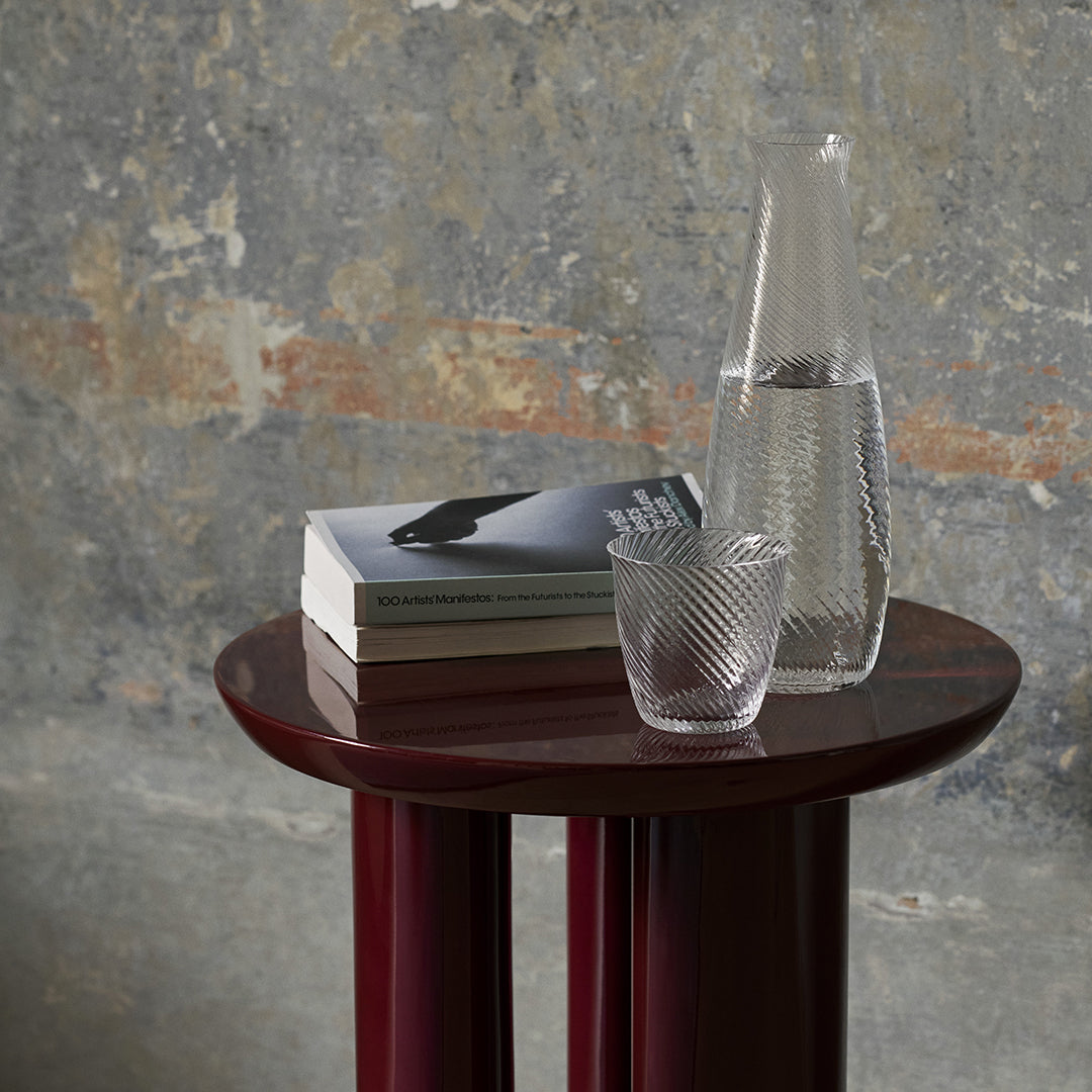 "Elegant maroon side table with textured glassware and patterned books against a textured gray wall backdrop."