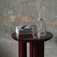 "Elegant maroon side table with textured glassware and patterned books against a textured gray wall backdrop."