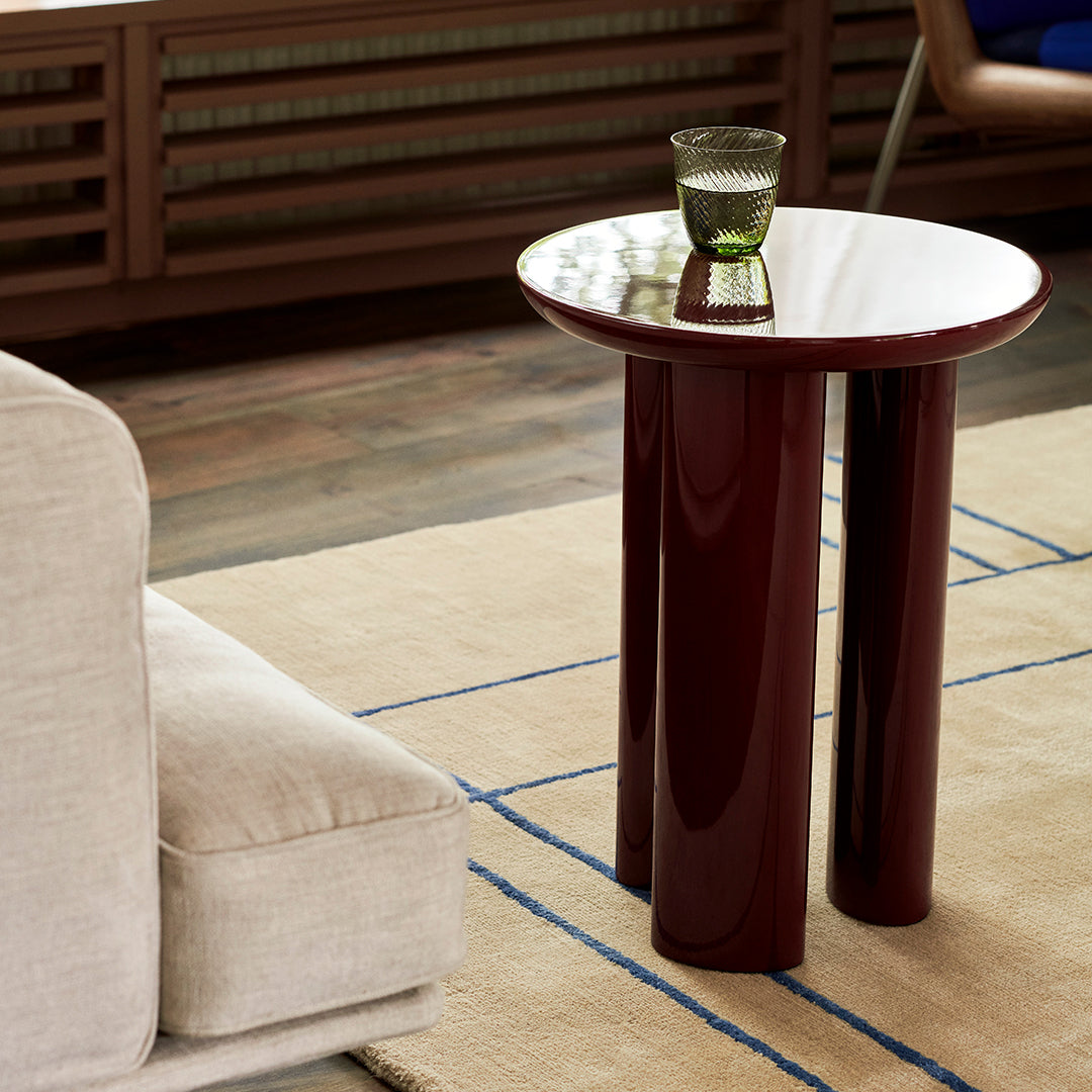 Round burgundy side table with glossy surface and three cylindrical legs, complemented by a clear glass on top.
