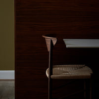 Elegant wooden chair with natural fiber seat against a rich dark wood wall and soft white table, highlighting warm tones.