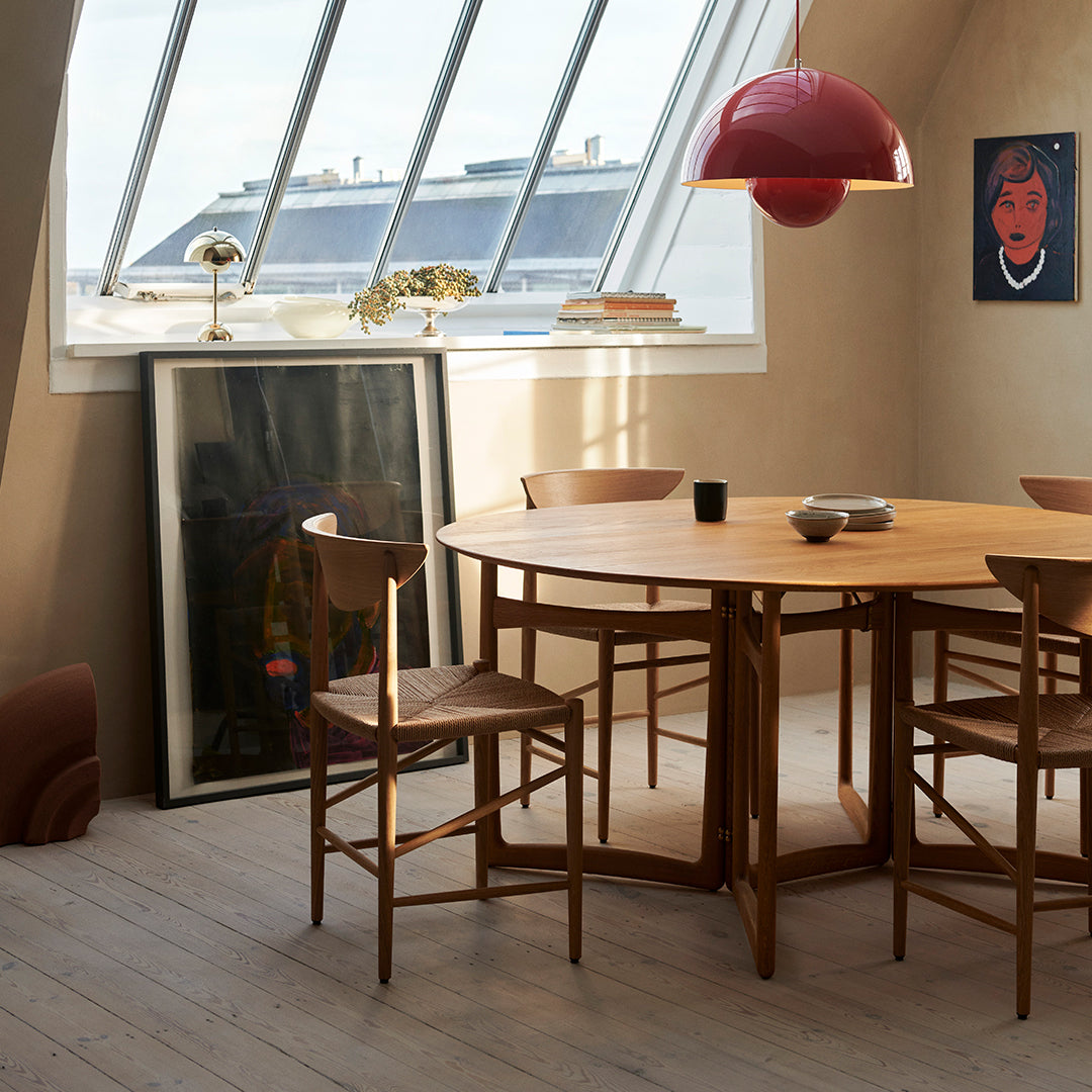 "Round wooden dining table with light brown finish, complemented by warm-toned chairs and a red pendant lamp."