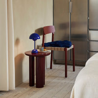 Red and blue chair and lamp with a round red table, set against a neutral wall and light wood floor.