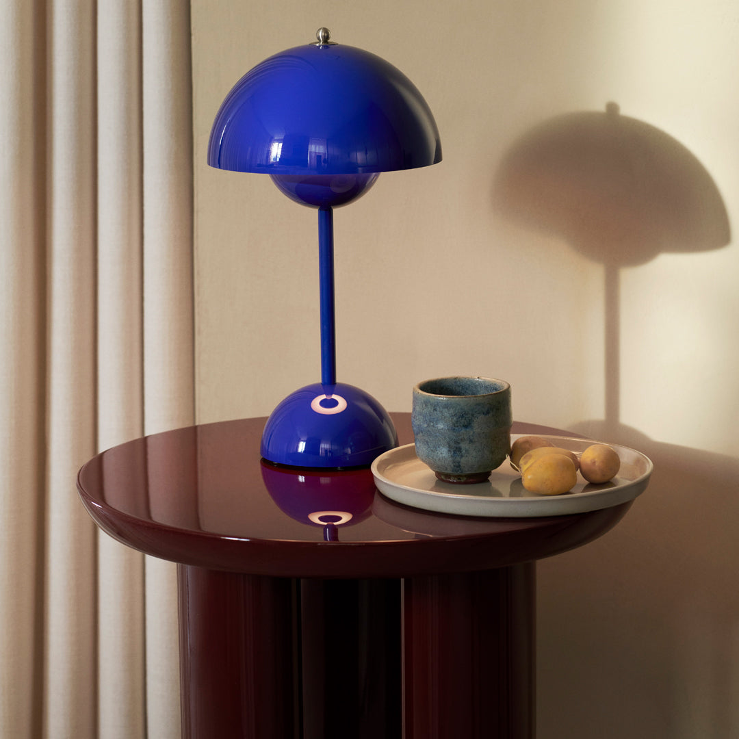 Vibrant blue lamp on a glossy maroon table, with a textured blue cup and neutral-toned plate displaying soft fruits.