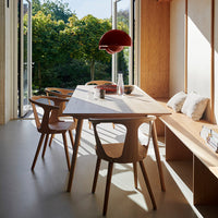 Wooden dining table with natural finish, paired with cream and red accents in a sunlit modern setting. Green trees outside.