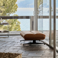 Tan leather ottoman with sleek black base, set against a modern stone floor and bright outdoor view.