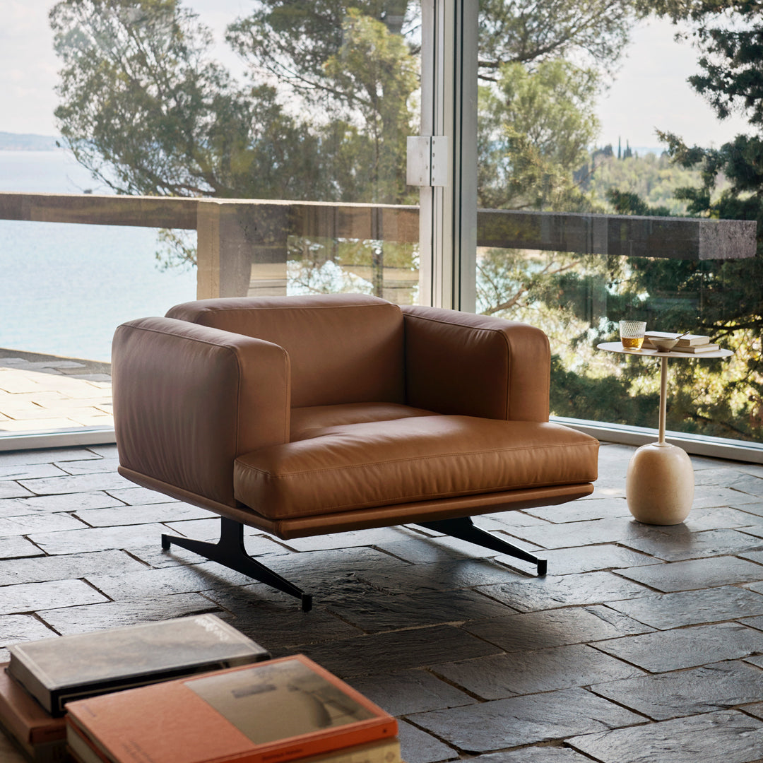 Brown leather armchair with minimalist design, resting on a sleek black base against a natural stone floor.