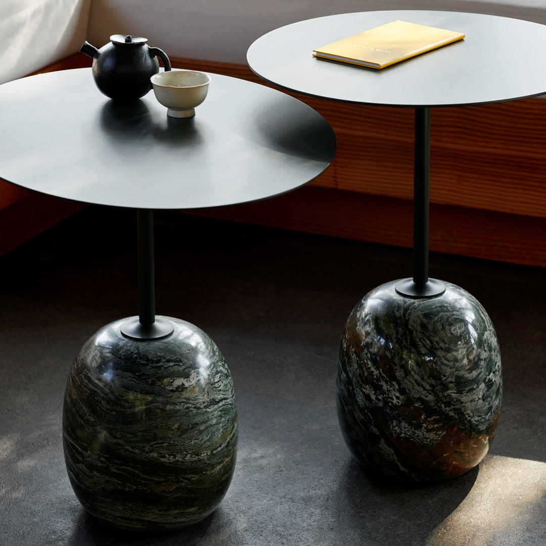 Modern round side tables with black metal tops and unique green, brown, and grey stone bases. Elegant texture and contrast.
