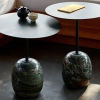 Modern round side tables with black metal tops and unique green, brown, and grey stone bases. Elegant texture and contrast.