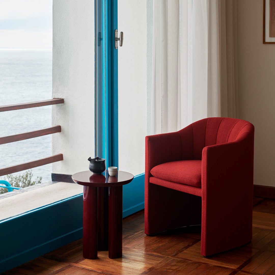 Red curved armchair next to a round dark table, with blue and white window frames in a bright, minimalistic setting.