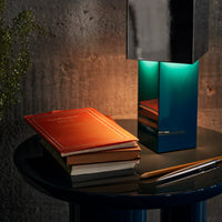 Modern lamp with a shiny black finish and turquoise glow, paired with stacked red and brown books on a sleek black table.