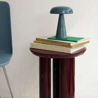 Stylish gray lamp on stacked books, featuring burgundy table surface, green accents, and soft blue chair.