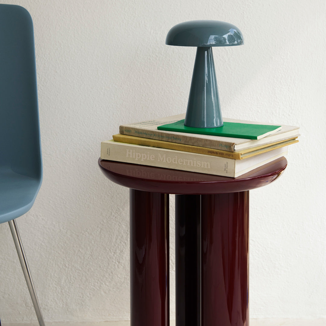 Stylish gray lamp on stacked books, featuring burgundy table surface, green accents, and soft blue chair.