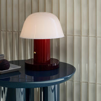 White dome lamp with a burgundy base on a blue table against a textured cream wall. Modern and sleek design.