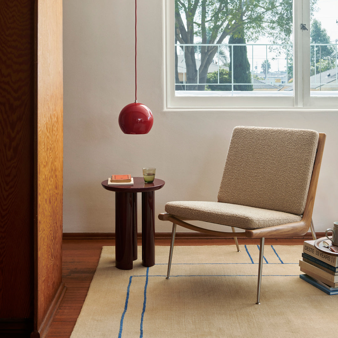 Beige upholstered chair with wooden legs, red pendant light, and a patterned rug with blue lines in a bright room.