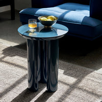 Glossy blue round side table with cylindrical legs, topped with a bowl of apples and a glass. Modern, sleek design.