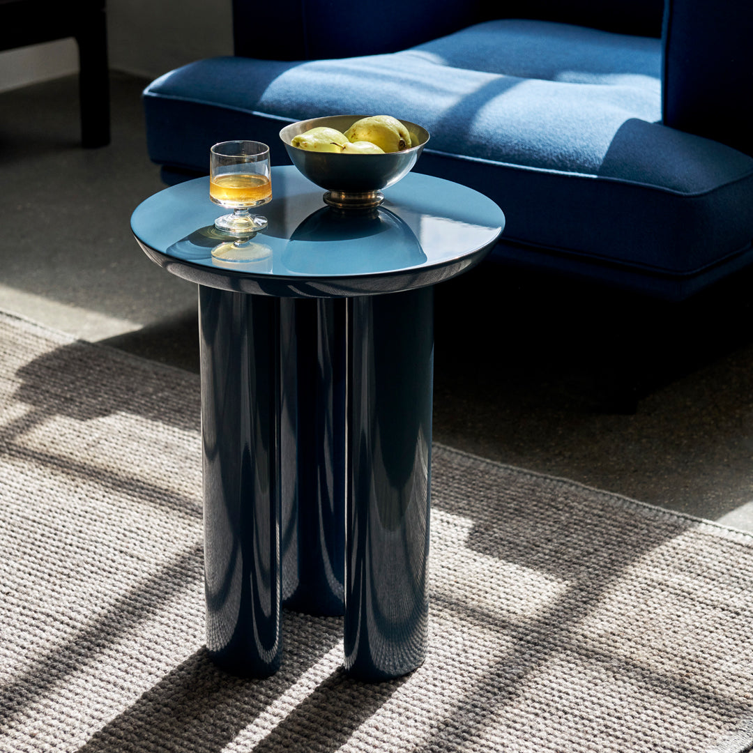 Glossy blue round side table with cylindrical legs, topped with a bowl of apples and a glass. Modern, sleek design.