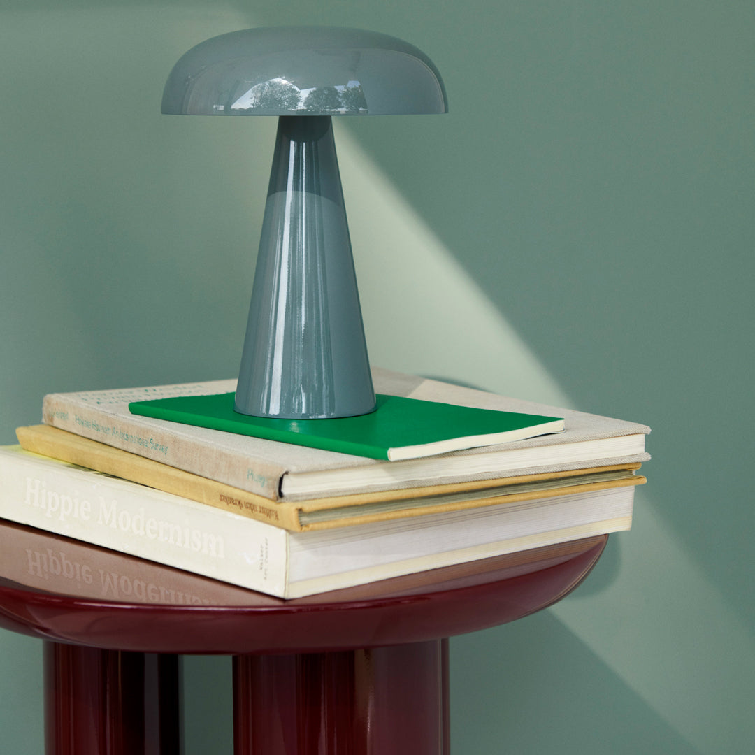 Modern lamp with a glossy gray finish atop stacked books with a green cover, against a muted green backdrop.