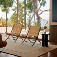 "Pair of natural woven chairs on a beige rug with subtle blue accents, set against a bright, green landscape."