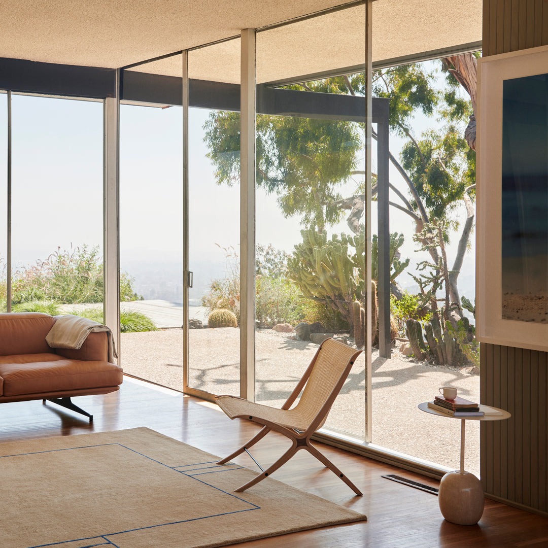 Bright, airy room featuring soft beige furniture, natural wood floors, and vibrant greenery through large glass windows.