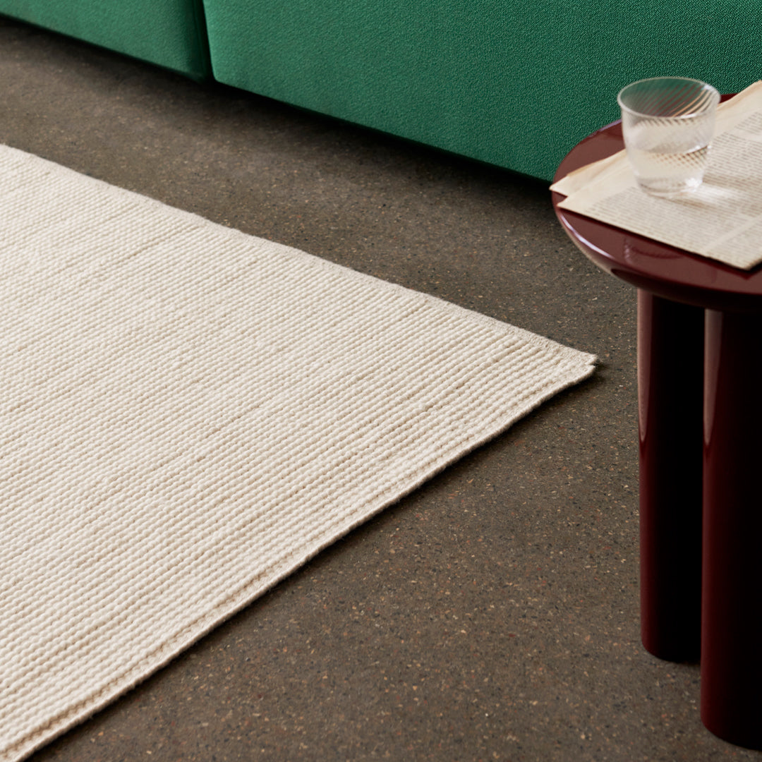 Textured cream area rug with subtle stripes, paired with a dark floor and green couch. Minimalist and modern design.