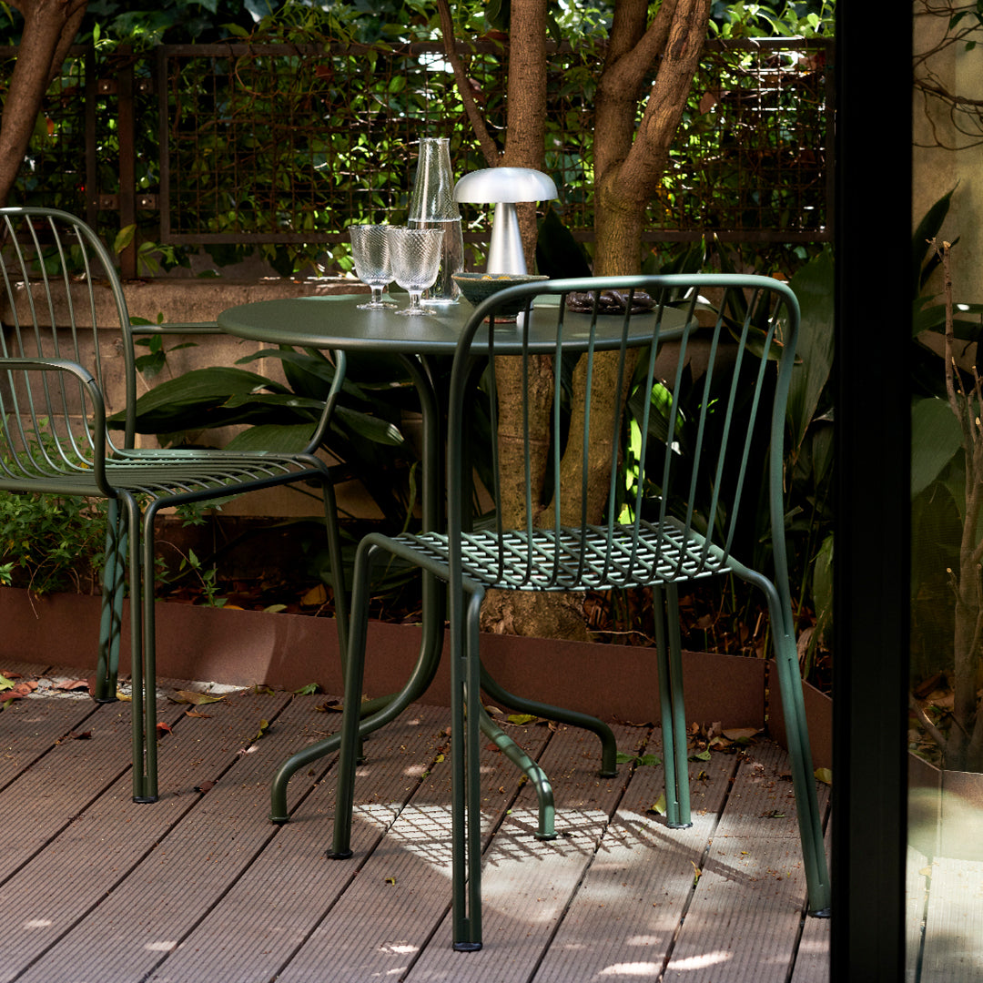 Green metal chairs with intricate patterns, complementing a round green table amidst lush greenery and wooden decking.