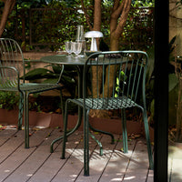 Green metal chairs with intricate patterns, complementing a round green table amidst lush greenery and wooden decking.
