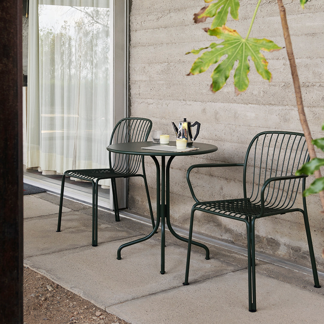 Green metal chairs and table with a minimalist design, set against a textured concrete wall and natural surroundings.