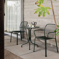 Green metal chairs and table with a minimalist design, set against a textured concrete wall and natural surroundings.