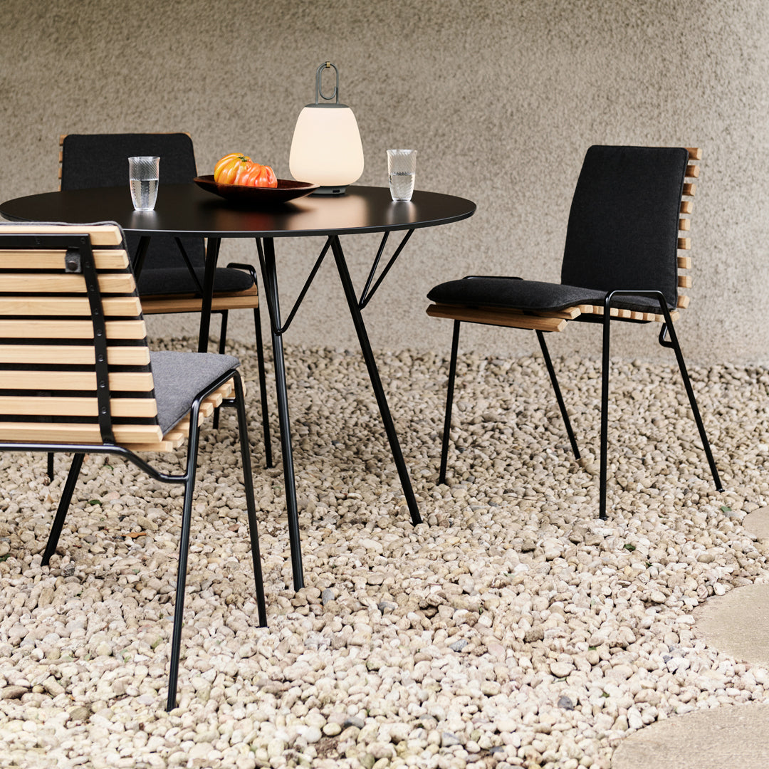 Round black table with sleek black chairs featuring wooden slats, set against a light gravel surface.