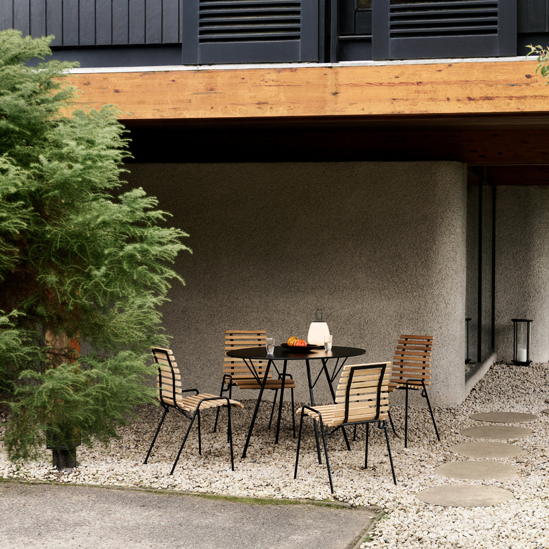 "Black metal frame dining set with light wood slats, set against a neutral stone wall and surrounded by greenery."
